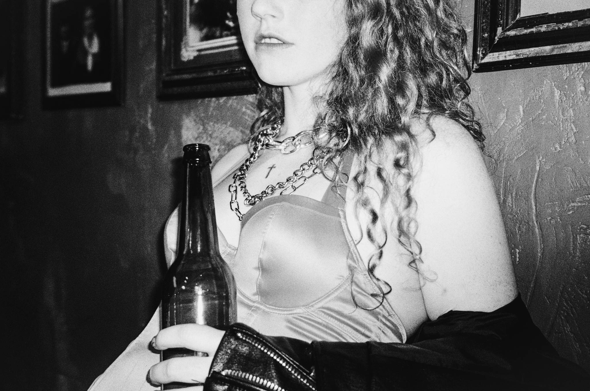 Black and white photo of a woman with curly hair holding a beer bottle indoors.