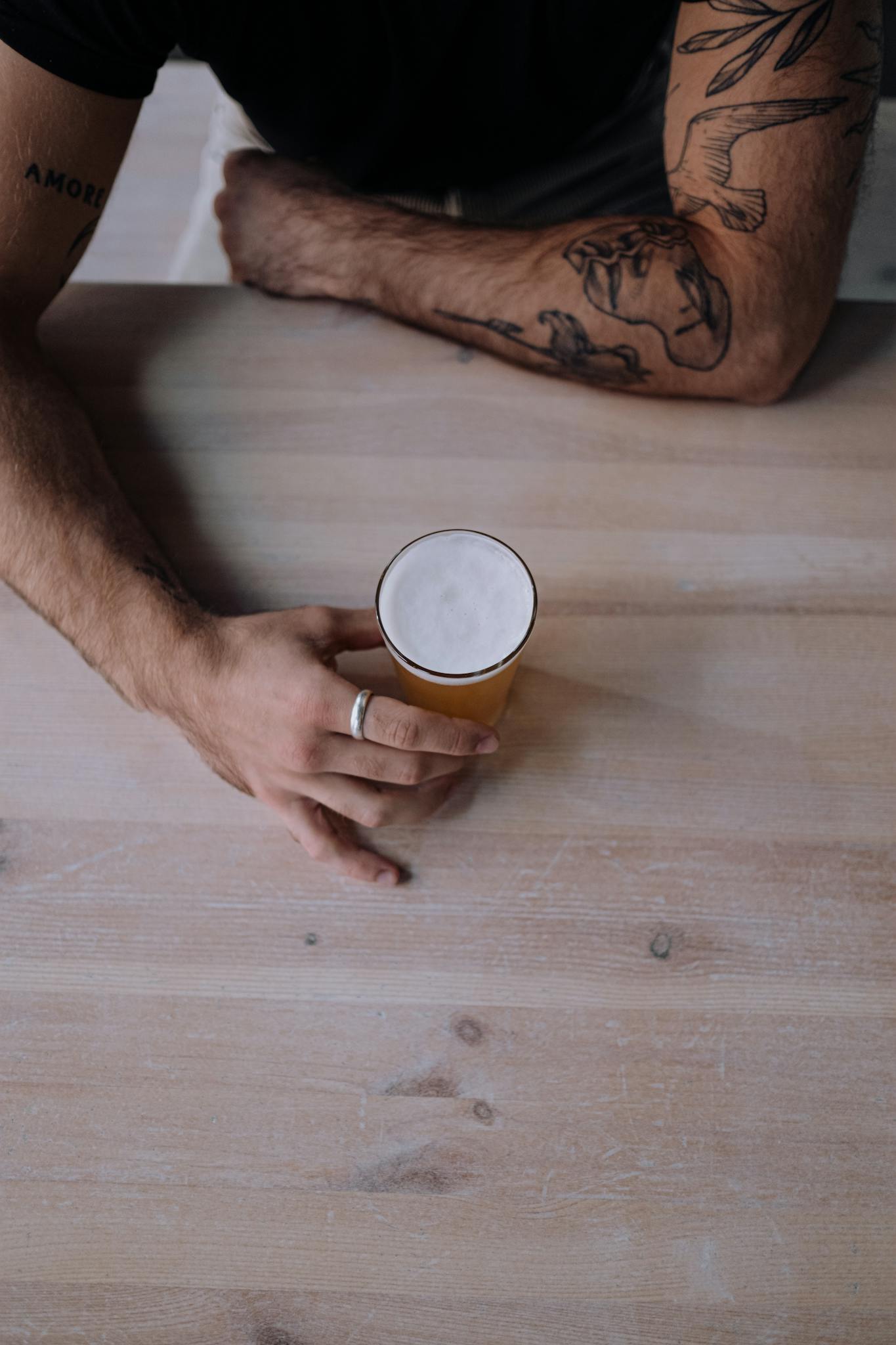 Overhead shot of a tattooed arm holding a craft beer on a rustic wooden table.