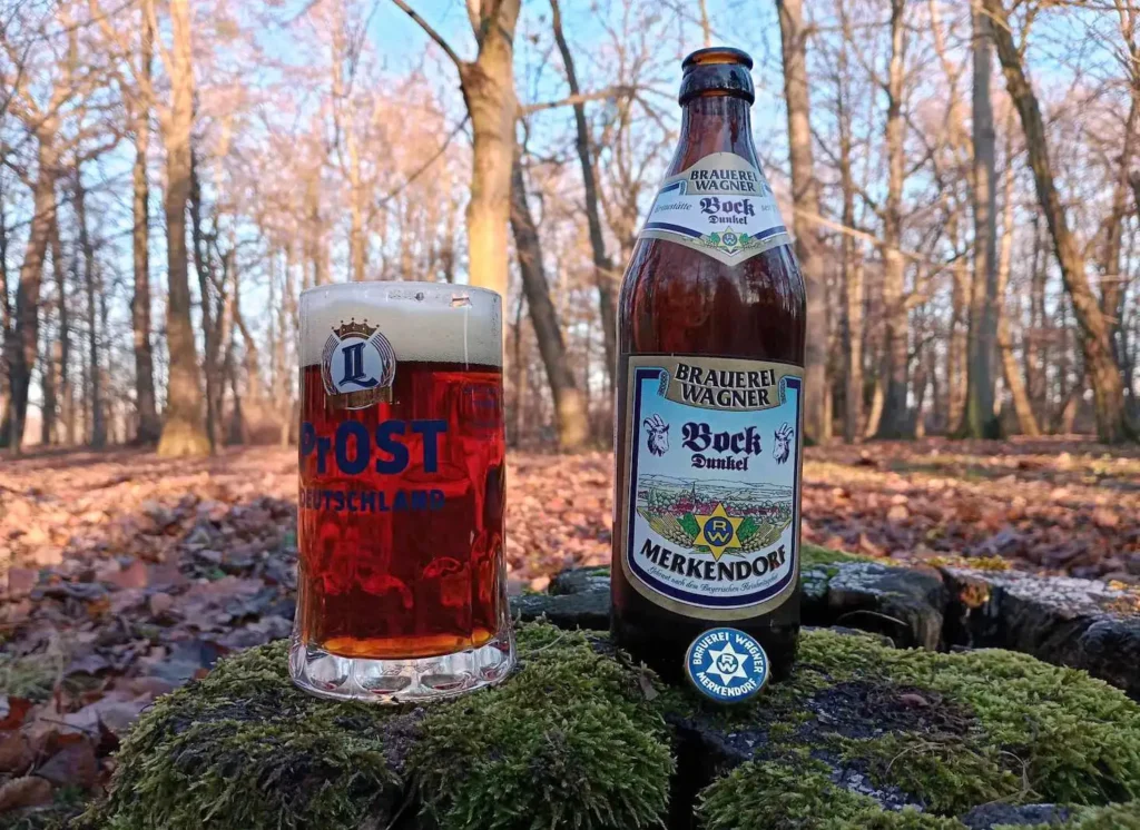Botella de Brauerei Wagner Bock Dunkel de Merkendorf junto a un vaso de cerveza alemán lleno con cerveza de color ámbar cobrizo oscuro y espuma cremosa, ambos posados sobre musgo en un entorno natural de bosque otoñal.
