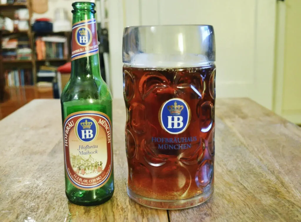 Botella Hofbräu Maibock con jarra tradicional bávara servida mostrando cerveza dorada cobriza, logo HB con corona real, ambiente casero con libros al fondo