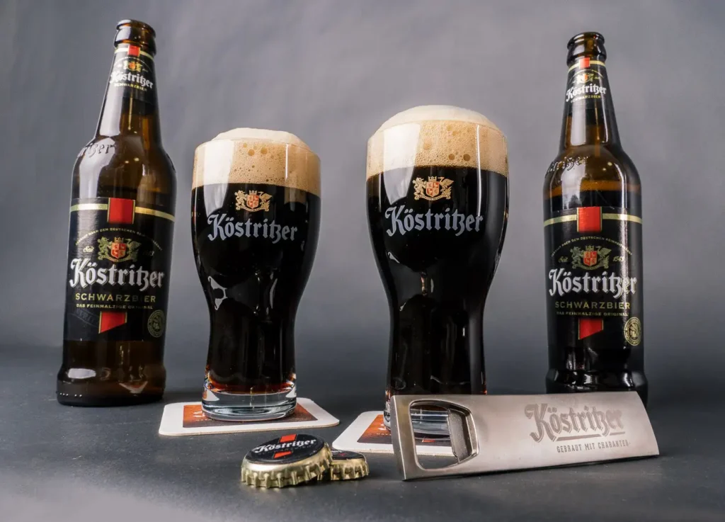 Botellas y vasos Köstritzer Schwarzbier alemana mostrando cerveza negra con espuma beige cremosa, abridor y posavasos de la marca histórica de Bad Köstritz