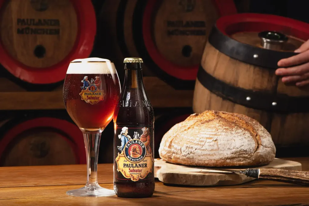 Botella Paulaner Salvator Doppelbock con copa servida mostrando cerveza marrón oscuro con reflejos cobrizos y espuma beige, pan artesanal y barriles de madera al fondo en bodega tradicional