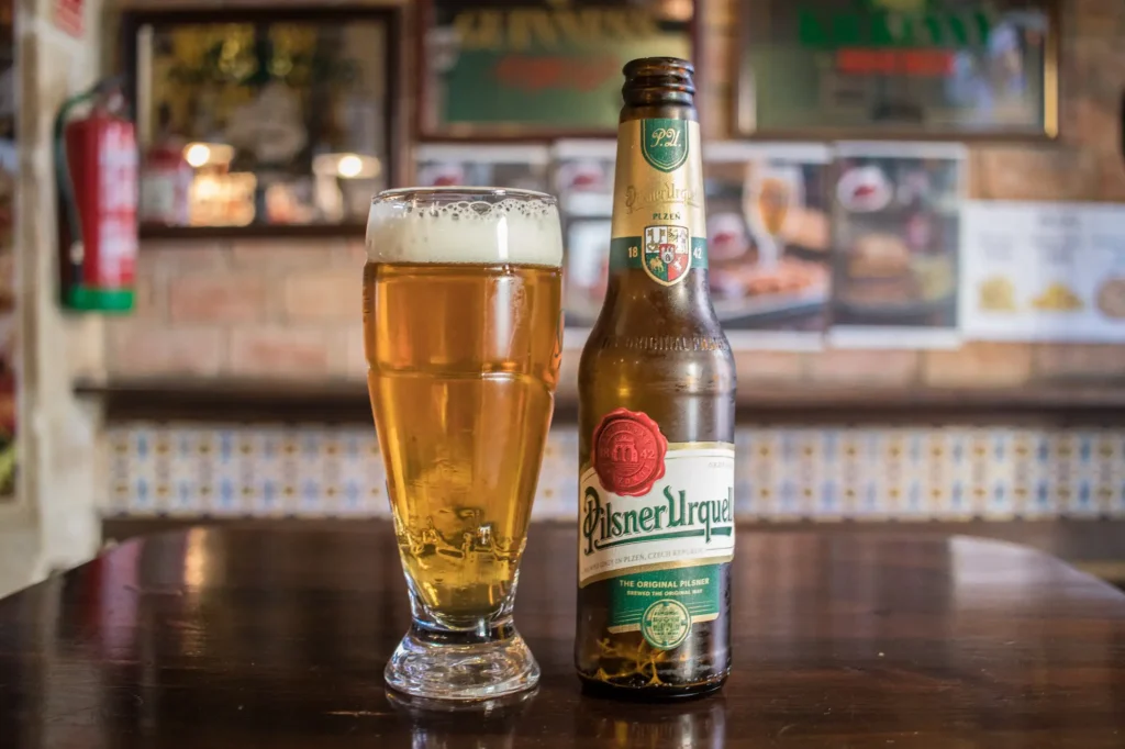 Botella de cerveza Pilsner Urquell junto a vaso servido con cerveza dorada y espuma sobre barra con azulejos decorativos al fondo, estética checa tradicional profesional