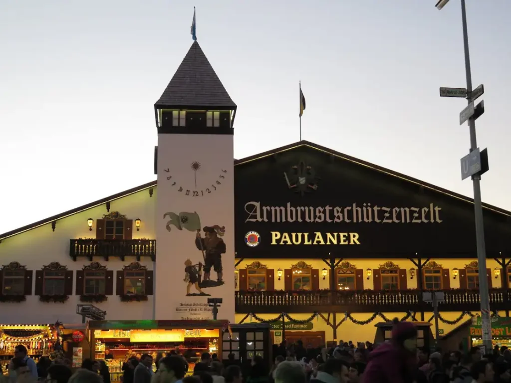 Fachada iluminada de la carpa Armbrustschützenzelt de Paulaner durante el Oktoberfest en Múnich, Alemania, al atardecer.
