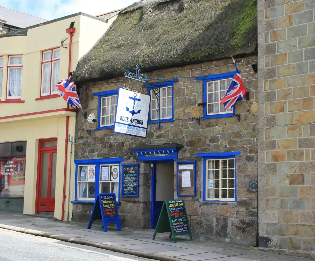 Fachada del histórico pub y cervecera Blue Anchor en Helston, Cornualles, Inglaterra, con banderas británicas y carteles en la entrada.
