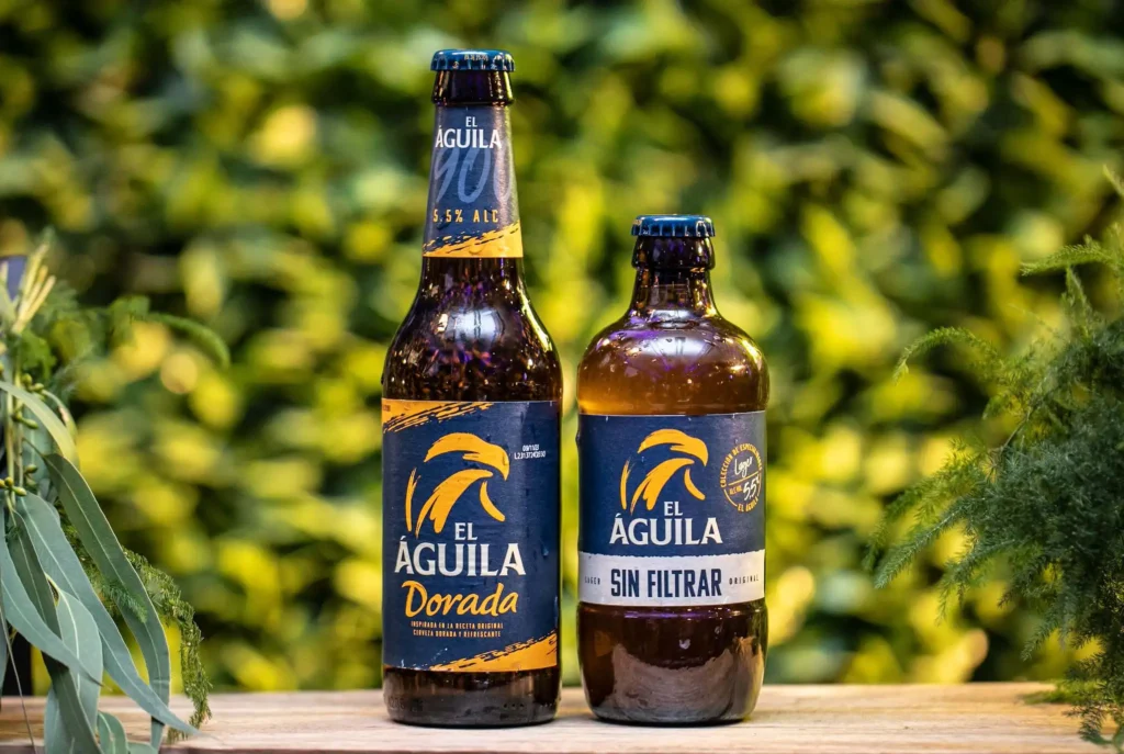 Botellas de cerveza El Águila Dorada y El Águila Sin Filtrar sobre una barra con el cartel luminoso "Saborea tu libertad" al fondo