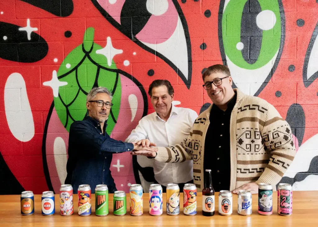Ignacio Rivera de Hijos de Rivera junto a los fundadores de Basqueland, Kevin Patricio y Ben Rozzi, posando frente a una colorida pared con sus cervezas artesanas en primer plano.